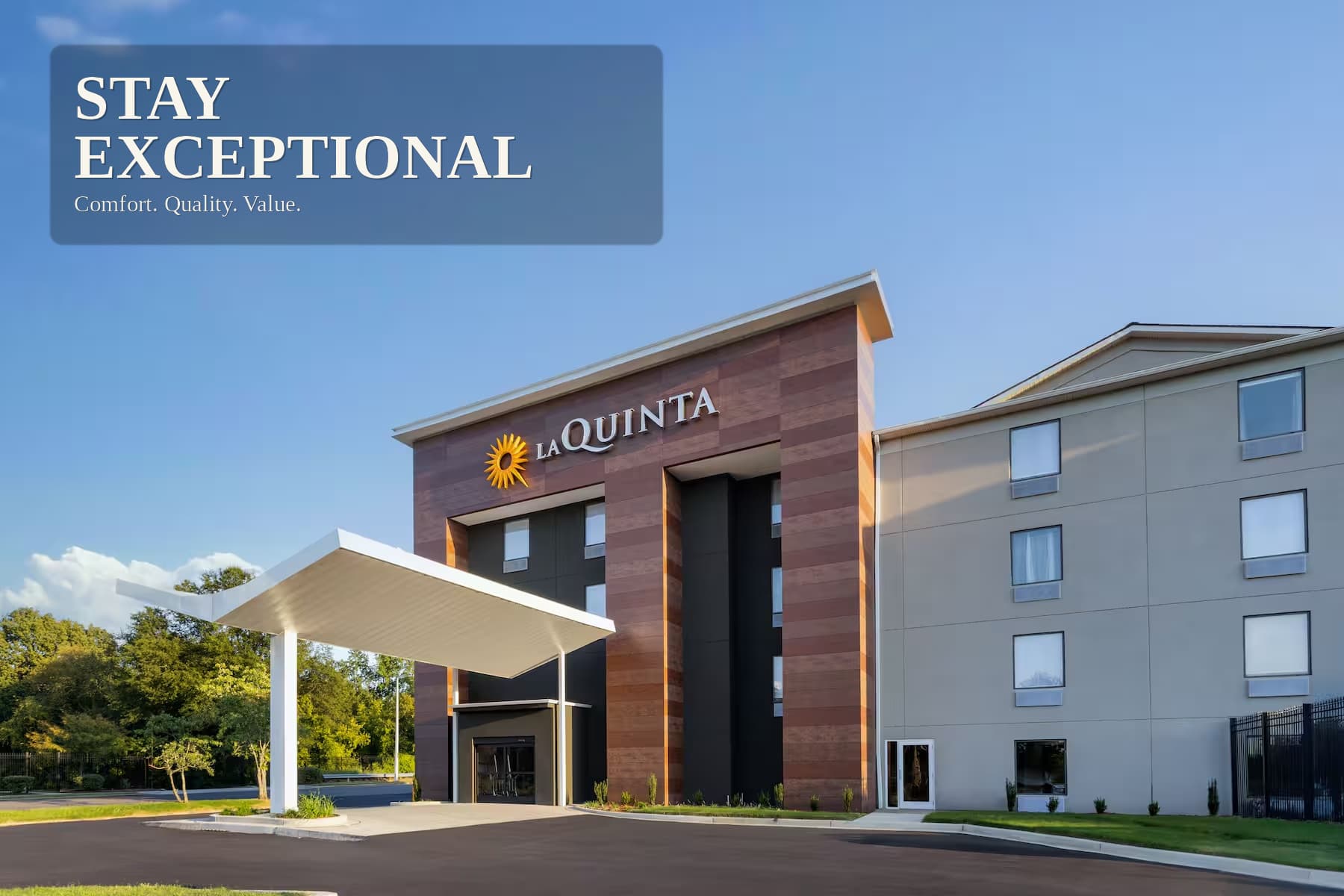 The hotel entrance in daylight — a timber-clad tower carrying the La Quinta by Wyndham sign, with the covered drive-through in front of it.