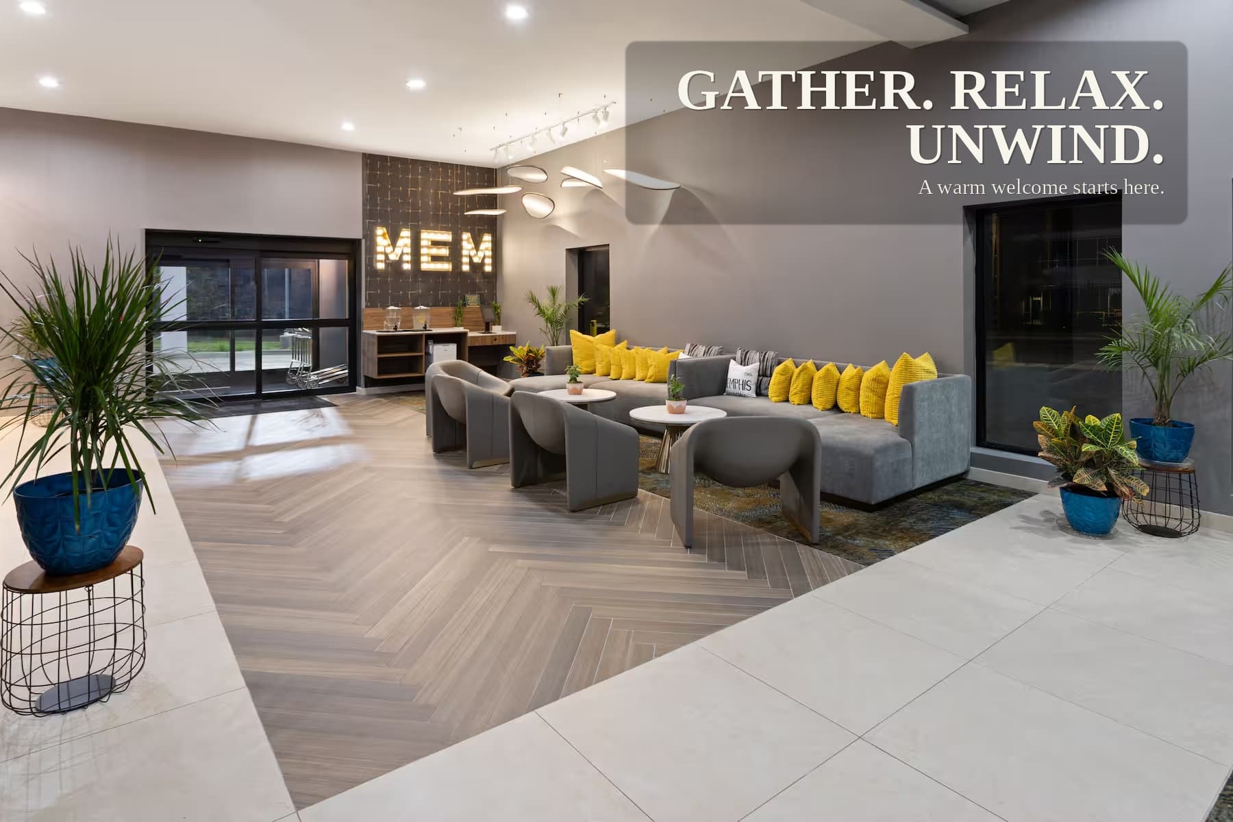 The lobby seating area: a long sectional with yellow cushions, low round tables, and a lit MEM marquee sign on the far wall beside the entrance doors.